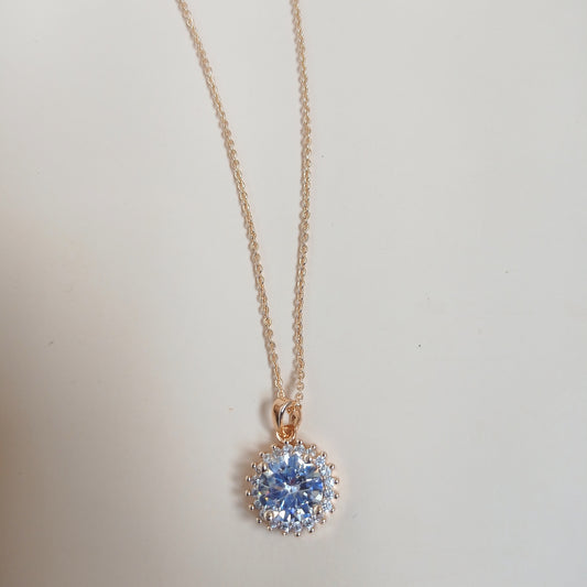 Elegant gold chain necklace featuring a dazzling round-cut blue gemstone pendant, surrounded by a halo of smaller stones, set against a neutral background