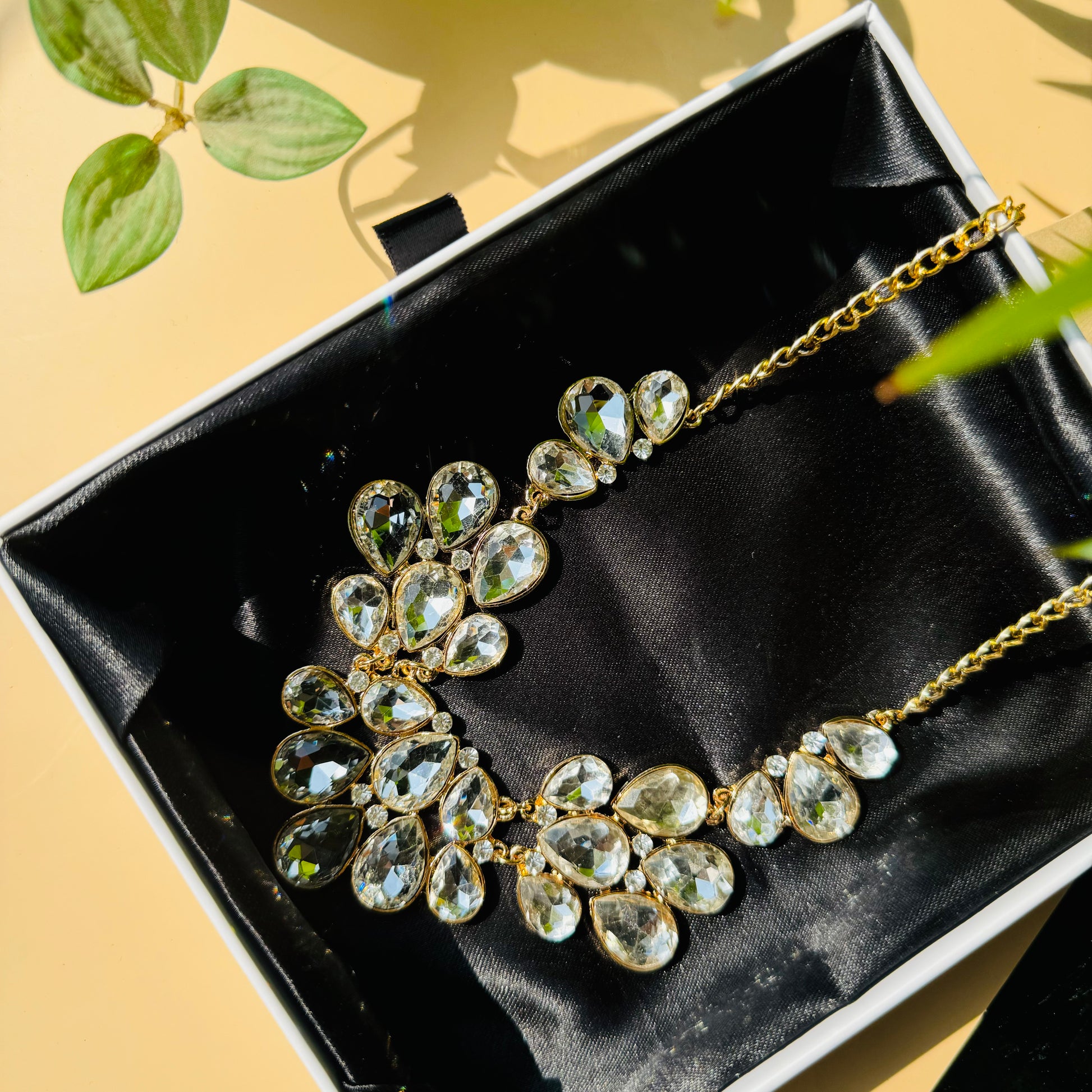 A stunning gold-tone statement necklace featuring an array of clear, faceted teardrop-shaped crystals arranged in a floral-inspired design, elegantly displayed on black satin fabric inside a jewelry box.