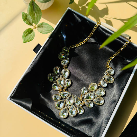 A stunning gold-tone statement necklace featuring an array of clear, faceted teardrop-shaped crystals arranged in a floral-inspired design, elegantly displayed on black satin fabric inside a jewelry box.