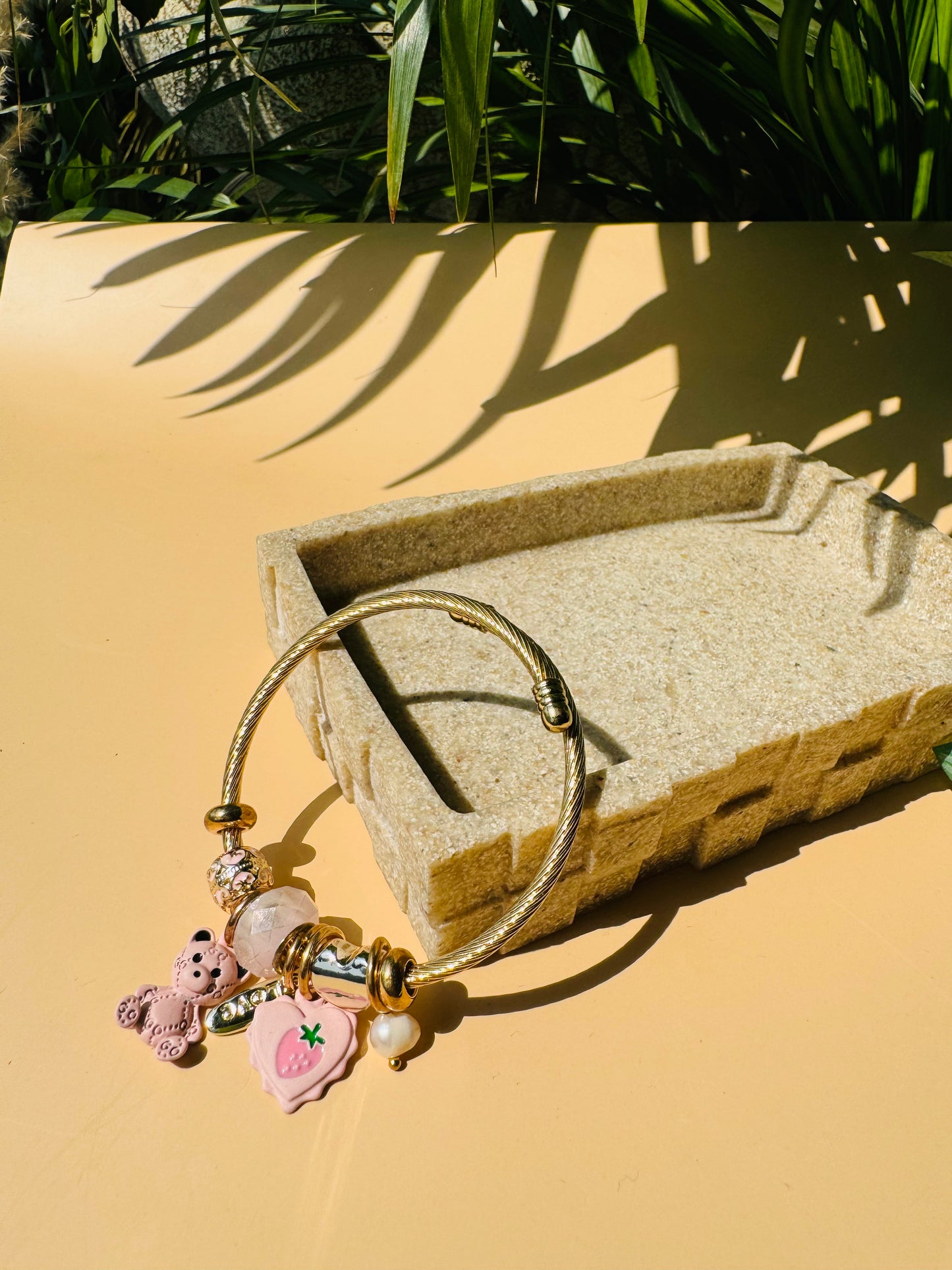 Elegant charm bracelet featuring a gold-colored flexible bangle adorned with various charms hearts and teddies. This Pandora-inspired bracelet is showcased on a neutral surface.