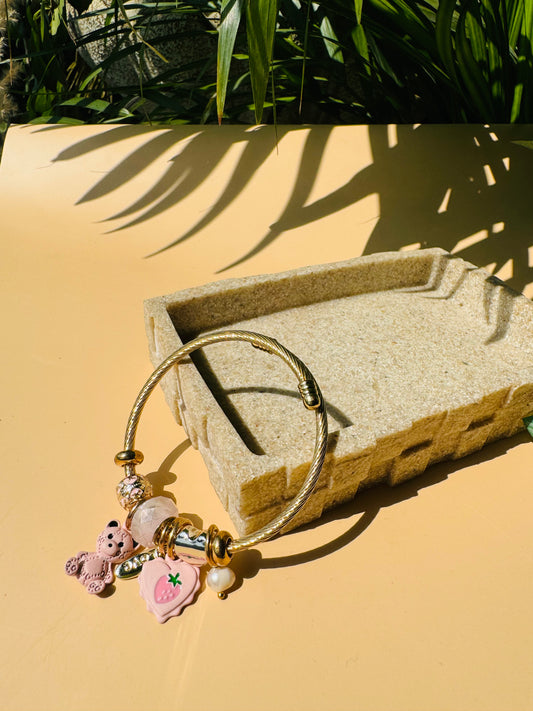 Elegant charm bracelet featuring a gold-colored flexible bangle adorned with various charms hearts and teddies. This Pandora-inspired bracelet is showcased on a neutral surface.
