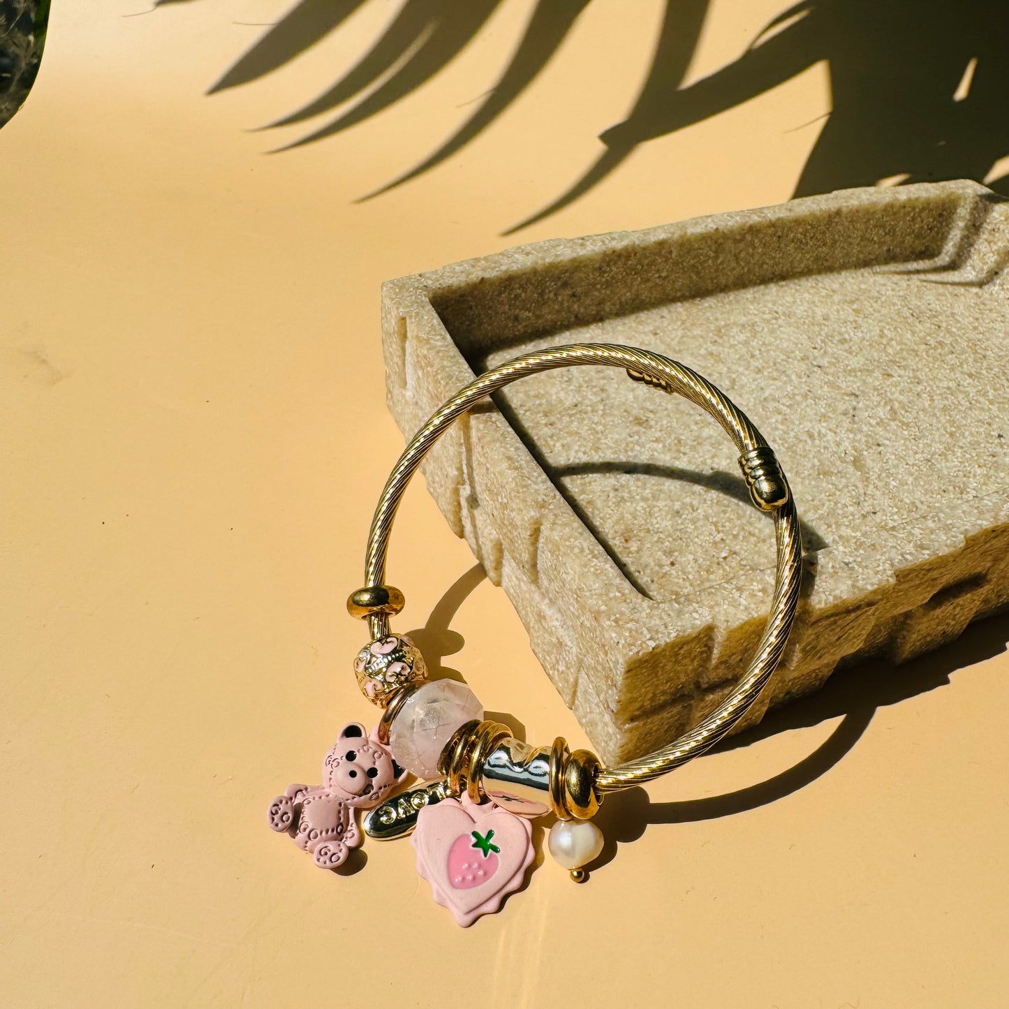 Elegant charm bracelet featuring a gold-colored flexible bangle adorned with various charms hearts and teddies. This Pandora-inspired bracelet is showcased on a neutral surface.