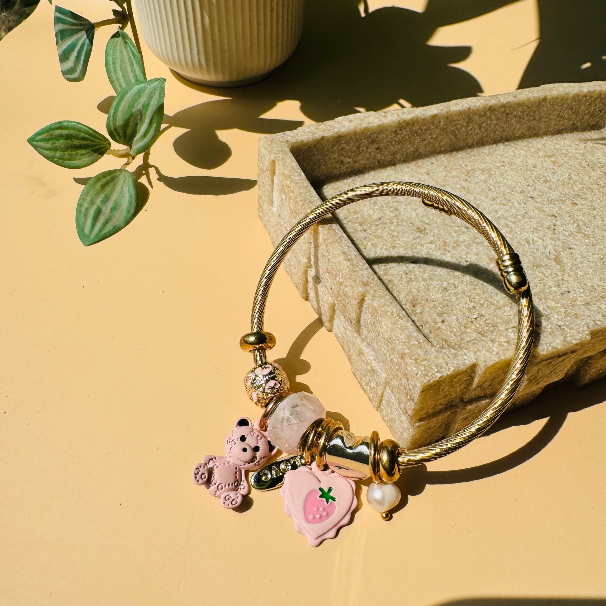 Elegant charm bracelet featuring a gold-colored flexible bangle adorned with various charms hearts and teddies. This Pandora-inspired bracelet is showcased on a neutral surface.