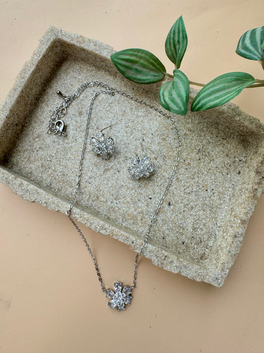 Elegant silver floral jewelry set featuring a delicate necklace with a matching pair of sparkling stud earrings, displayed on a textured beige tray with green leaf accents.