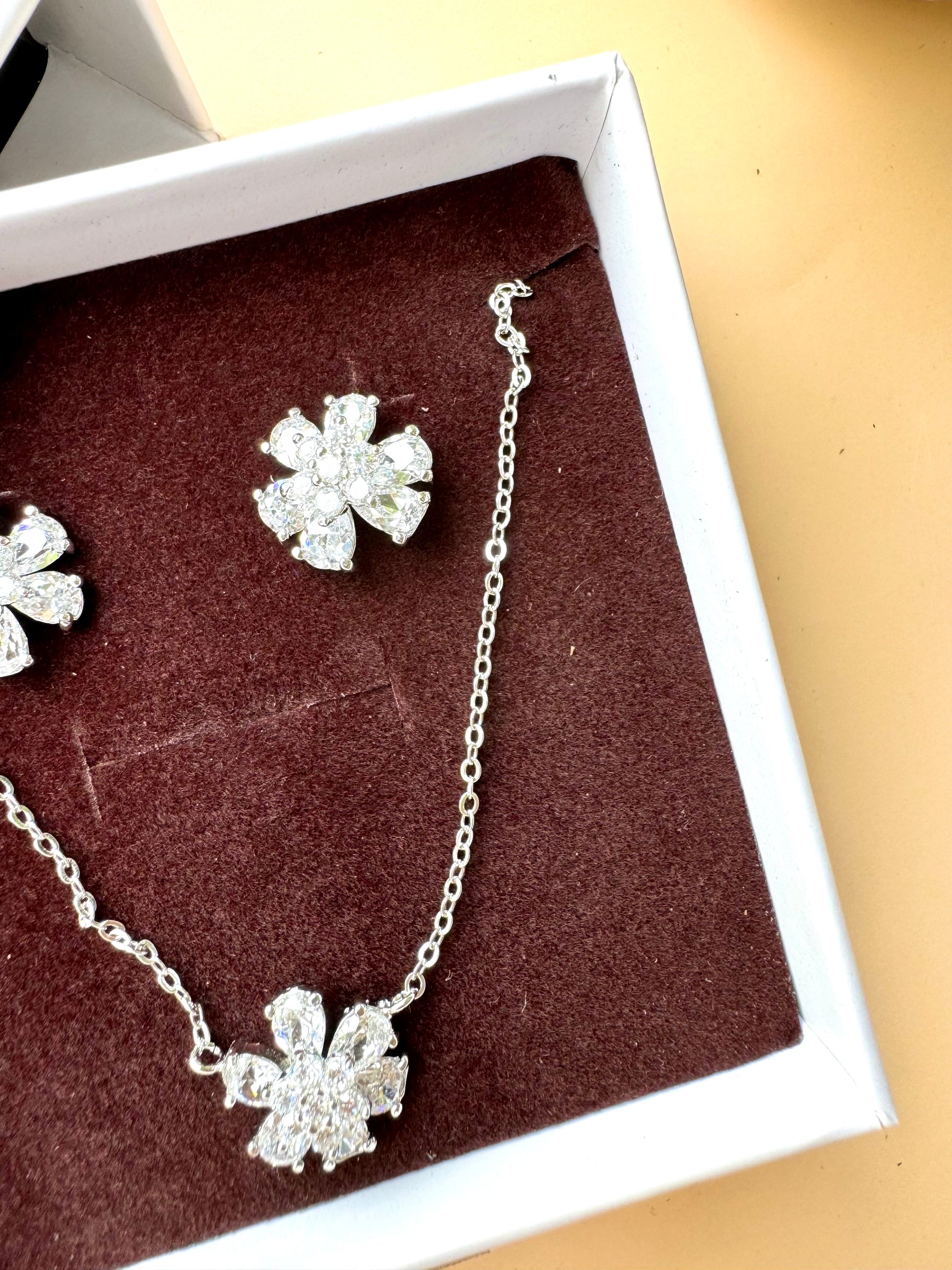 Elegant silver floral jewelry set featuring a delicate necklace with a matching pair of sparkling stud earrings, displayed on a textured beige tray with green leaf accents.