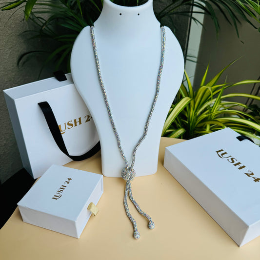 Alt text: A sparkling crystal lariat necklace displayed on a white bust stand. The necklace features a long, shimmering chain with a central crystal ball that secures two dangling strands, each ending in a smaller crystal-encrusted bead. The background includes Lush 24 branded packaging and lush green plants.