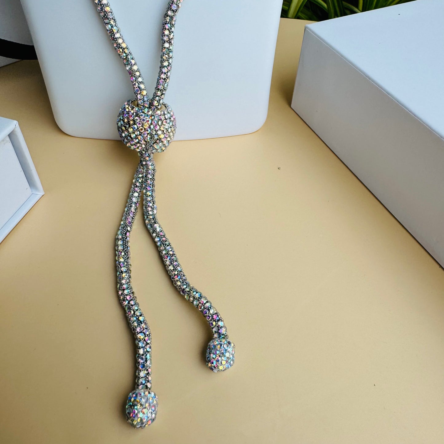 Alt text: A sparkling crystal lariat necklace displayed on a white bust stand. The necklace features a long, shimmering chain with a central crystal ball that secures two dangling strands, each ending in a smaller crystal-encrusted bead. The background includes Lush 24 branded packaging and lush green plants.