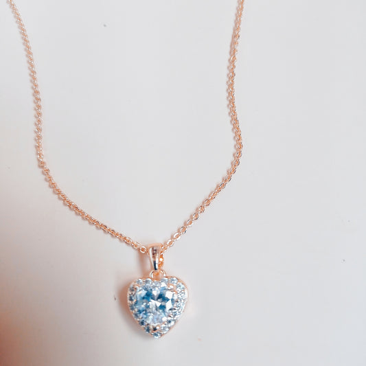 Rose gold necklace with a heart-shaped blue gemstone pendant, surrounded by sparkling white stones, set against a neutral background