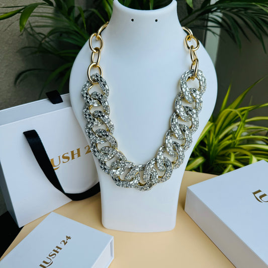 A bold, chunky statement necklace featuring oversized gold-linked loops encrusted with shimmering diamante crystals, displayed on a white bust against a stylish background with LUSH 24 packaging.