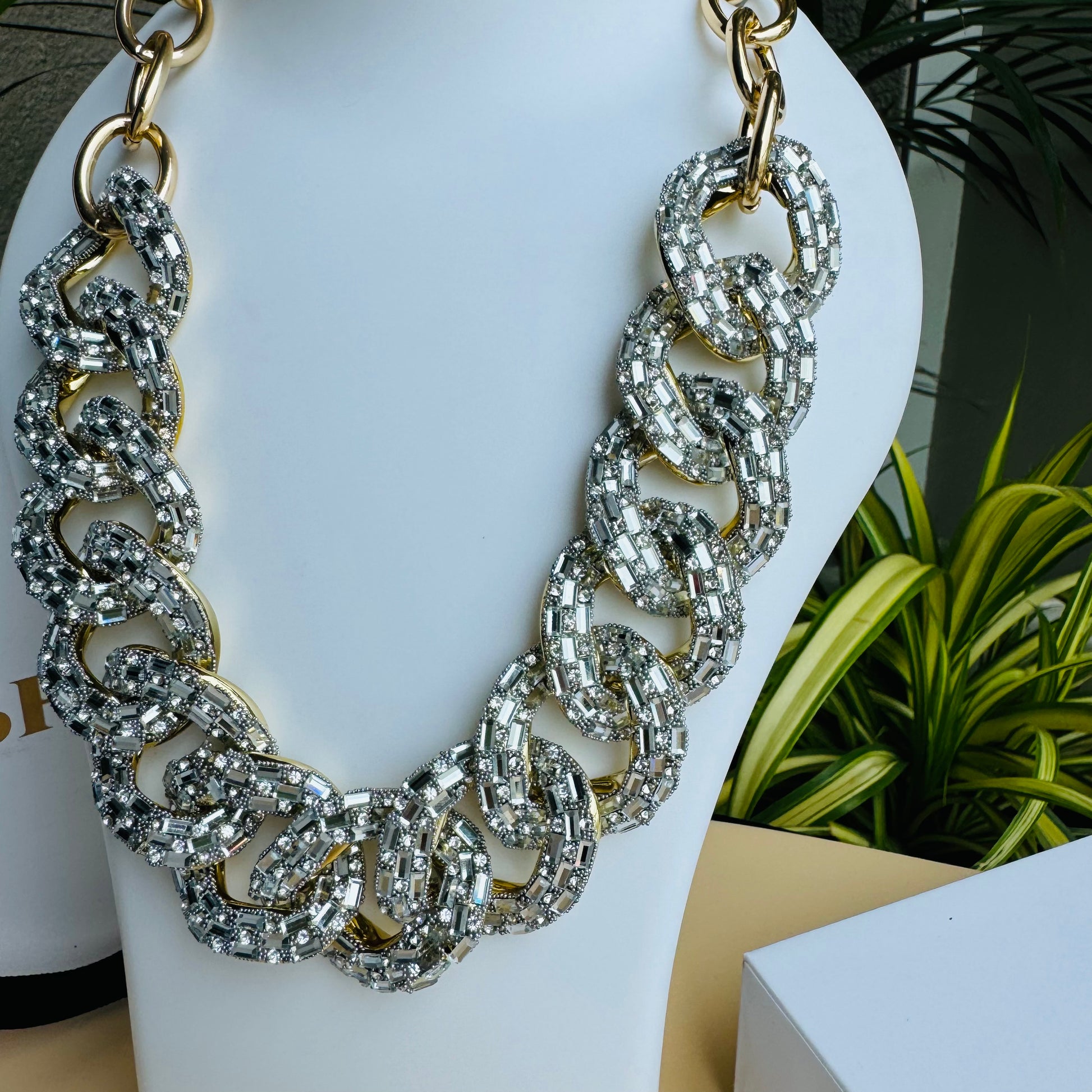 A bold, chunky statement necklace featuring oversized gold-linked loops encrusted with shimmering diamante crystals, displayed on a white bust against a stylish background with LUSH 24 packaging.
