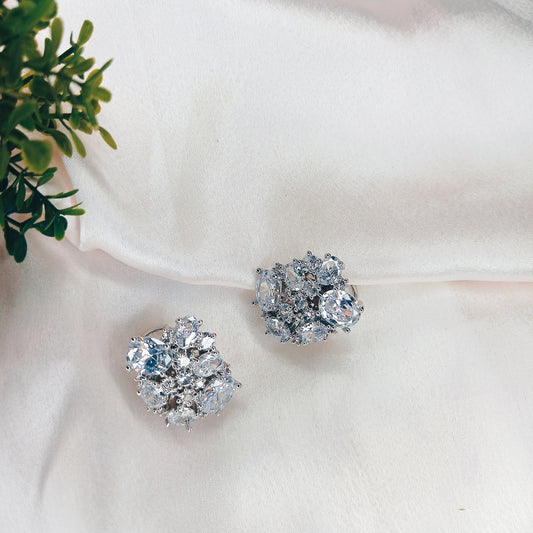 Elegant silver-tone floral cluster earrings adorned with sparkling cubic zirconia stones, arranged in a delicate, intricate design. Set against a soft, white satin background, these statement earrings exude luxury and sophistication, perfect for special occasions.