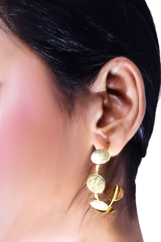 a woman wearing contemporary gold-tone statement earrings featuring multiple brushed metal discs in an artistic, asymmetric arrangement. The earrings have a modern, elegant look, perfect for adding a touch of sophistication to any outfit.