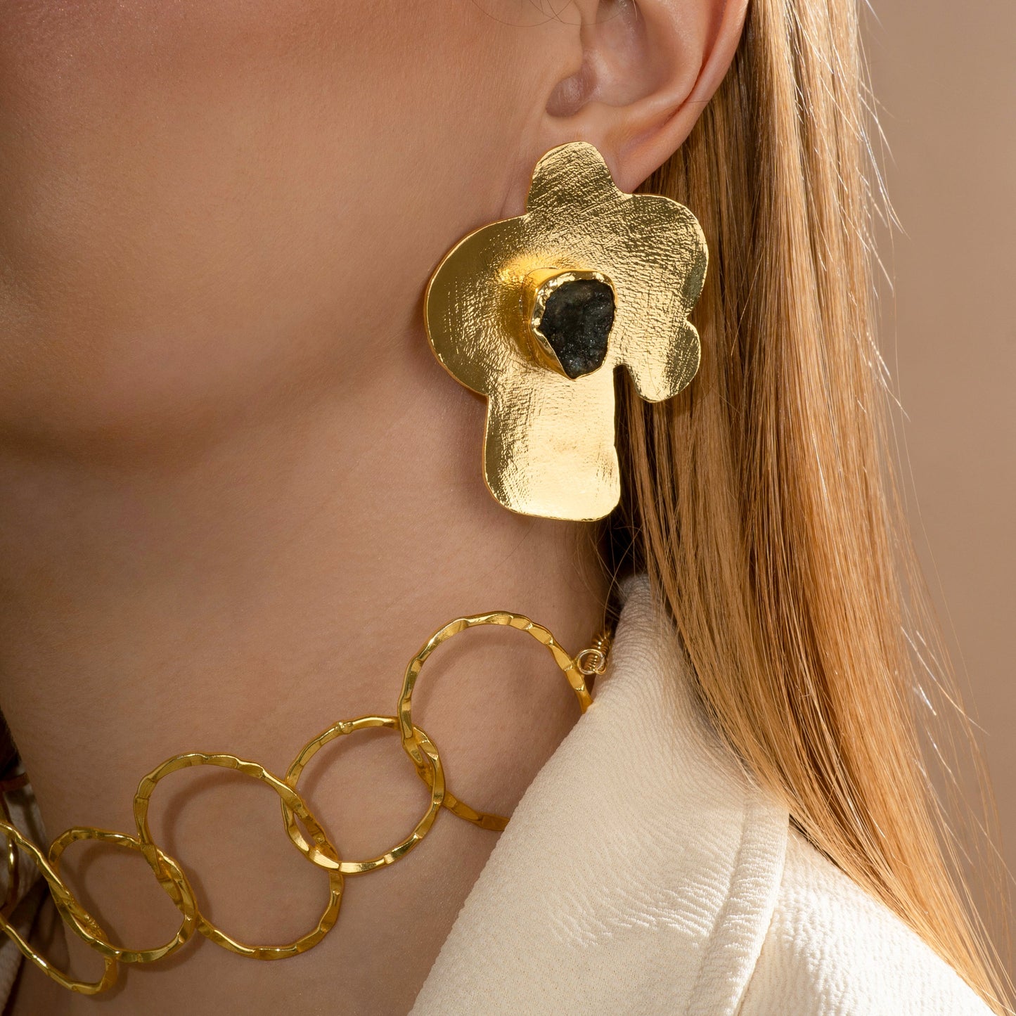Model wearing a statement gold-toned abstract floral earring with a black gemstone detail, paired with a gold chain necklace