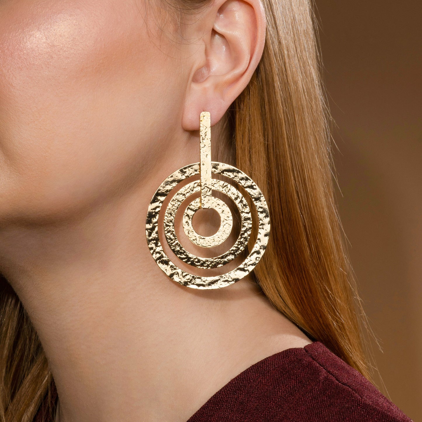 Close-up of a model wearing a gold-toned circular stud earring with a concentric spiral design, set against a dark background