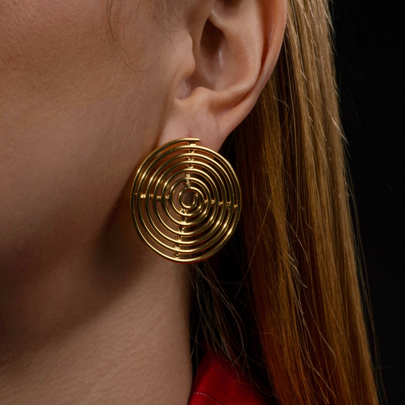 Close-up of a woman's ear adorned with a bold gold spiral statement earring featuring a concentric circular design, set against a dark background.