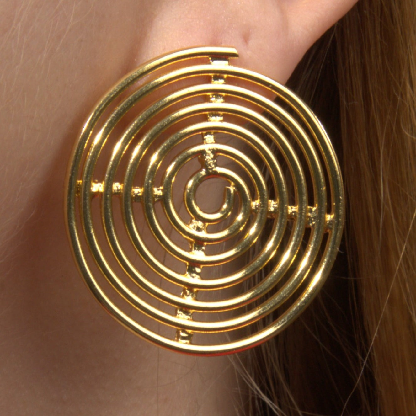 Close-up of a woman's ear adorned with a bold gold spiral statement earring featuring a concentric circular design, set against a dark background.
