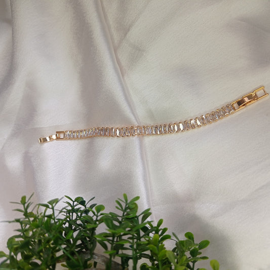 Elegant gold bracelet with baguette-cut clear gemstones displayed on a soft white satin fabric, with a touch of greenery in the foreground