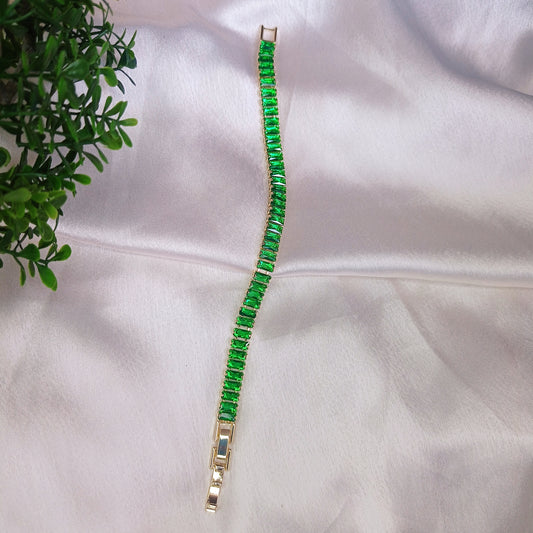 Elegant gold bracelet with vibrant green baguette-cut gemstones, displayed on soft satin fabric with a touch of greenery