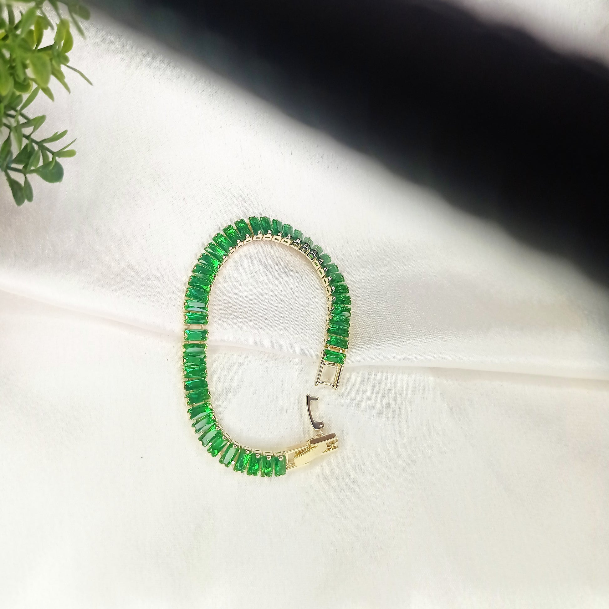 Elegant gold bracelet with vibrant green baguette-cut gemstones, displayed on soft satin fabric with a touch of greenery