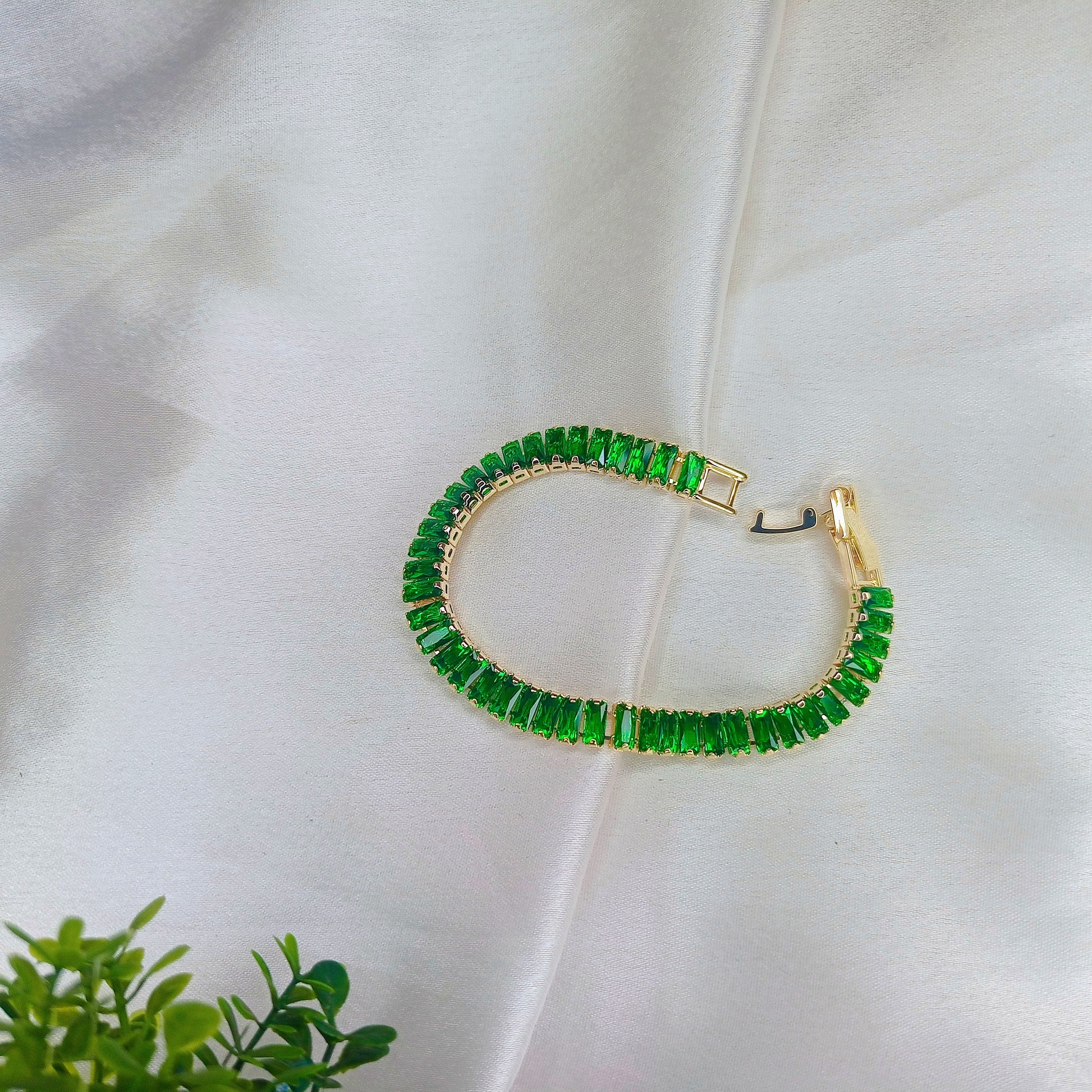 Elegant gold bracelet with vibrant green baguette-cut gemstones, displayed on soft satin fabric with a touch of greenery