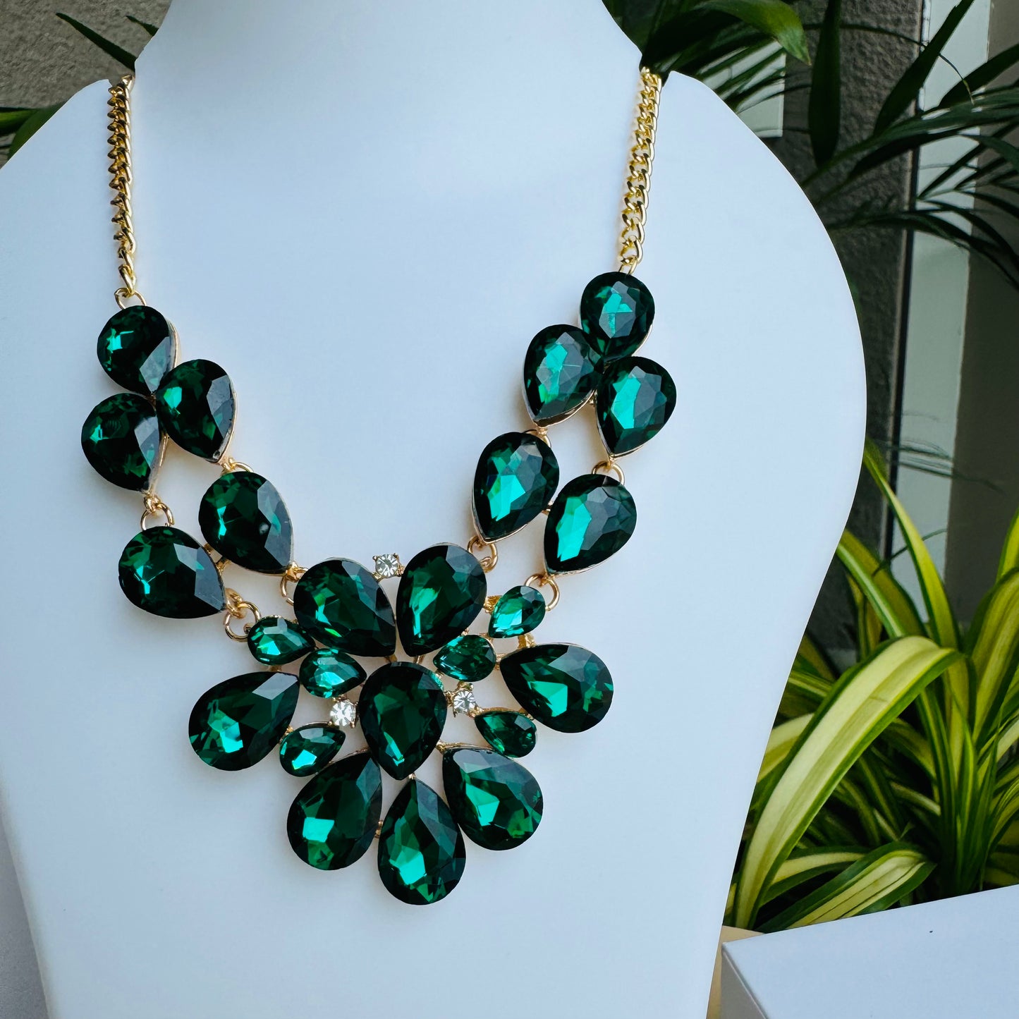 Statement necklace with faceted emerald green and black gemstones on a gold chain, displayed on a white mannequin bust. The intricate design features teardrop and oval-cut stones, creating a bold and luxurious look.