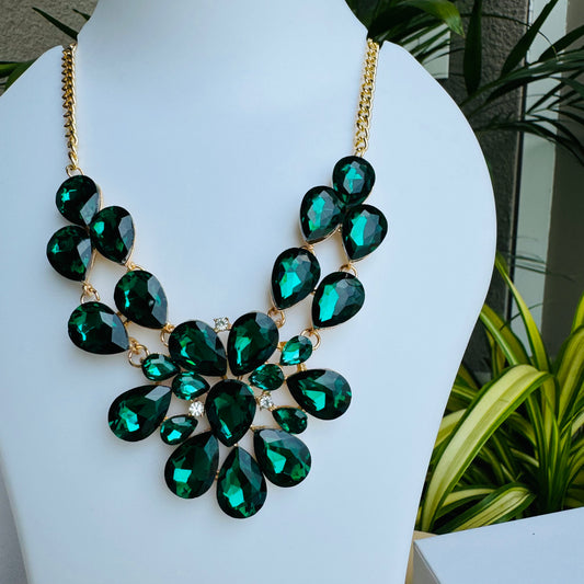 Statement necklace with faceted emerald green and black gemstones on a gold chain, displayed on a white mannequin bust. The intricate design features teardrop and oval-cut stones, creating a bold and luxurious look.