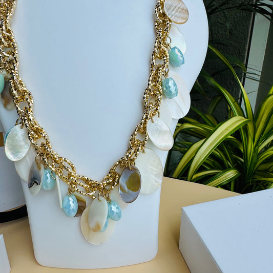 Gold-tone chunky necklace adorned with enamel-finished shell discs and faceted blue beads, displayed on a white bust stand against a backdrop of green plants and luxury packaging.