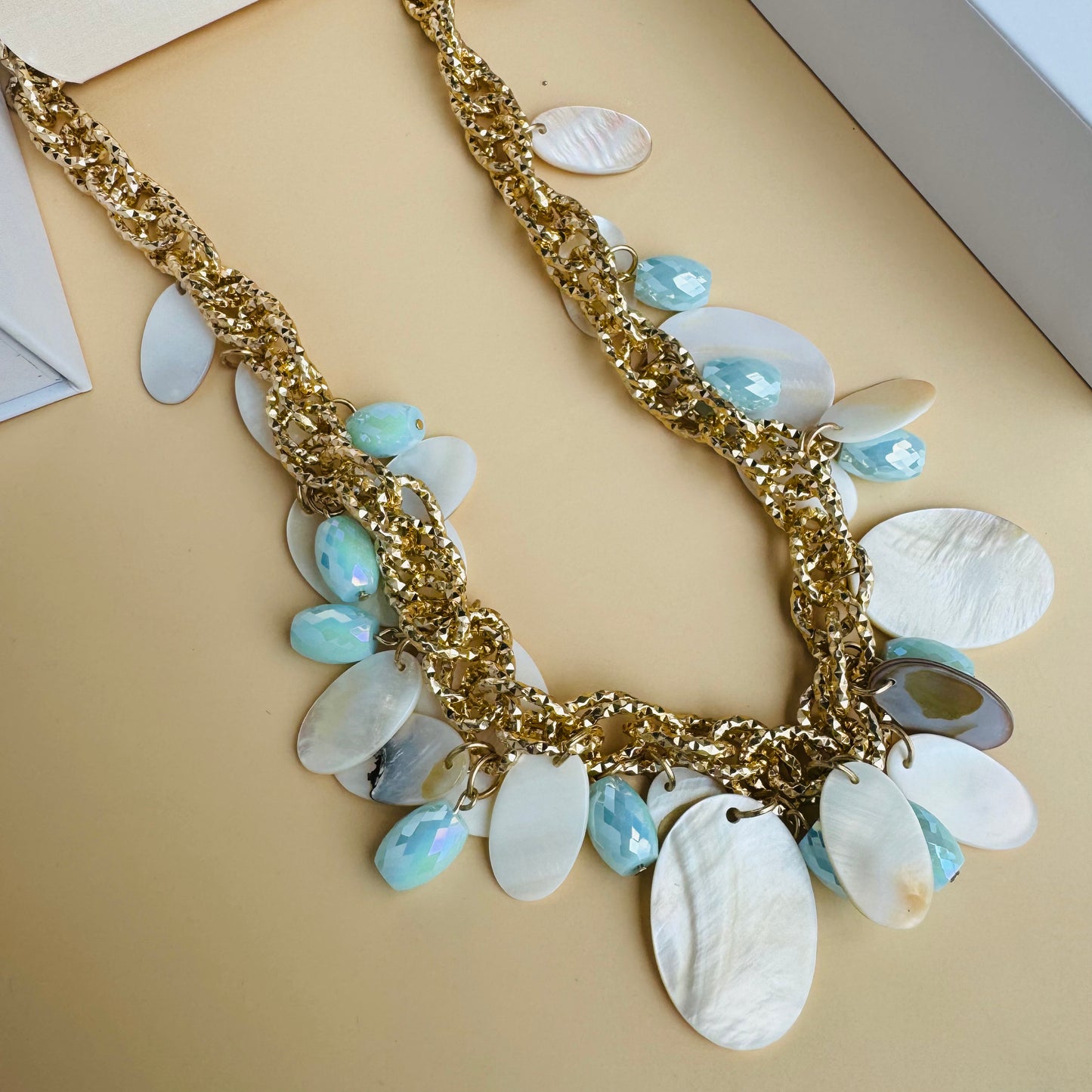 Gold-tone chunky necklace adorned with enamel-finished shell discs and faceted blue beads, displayed on a white bust stand against a backdrop of green plants and luxury packaging.