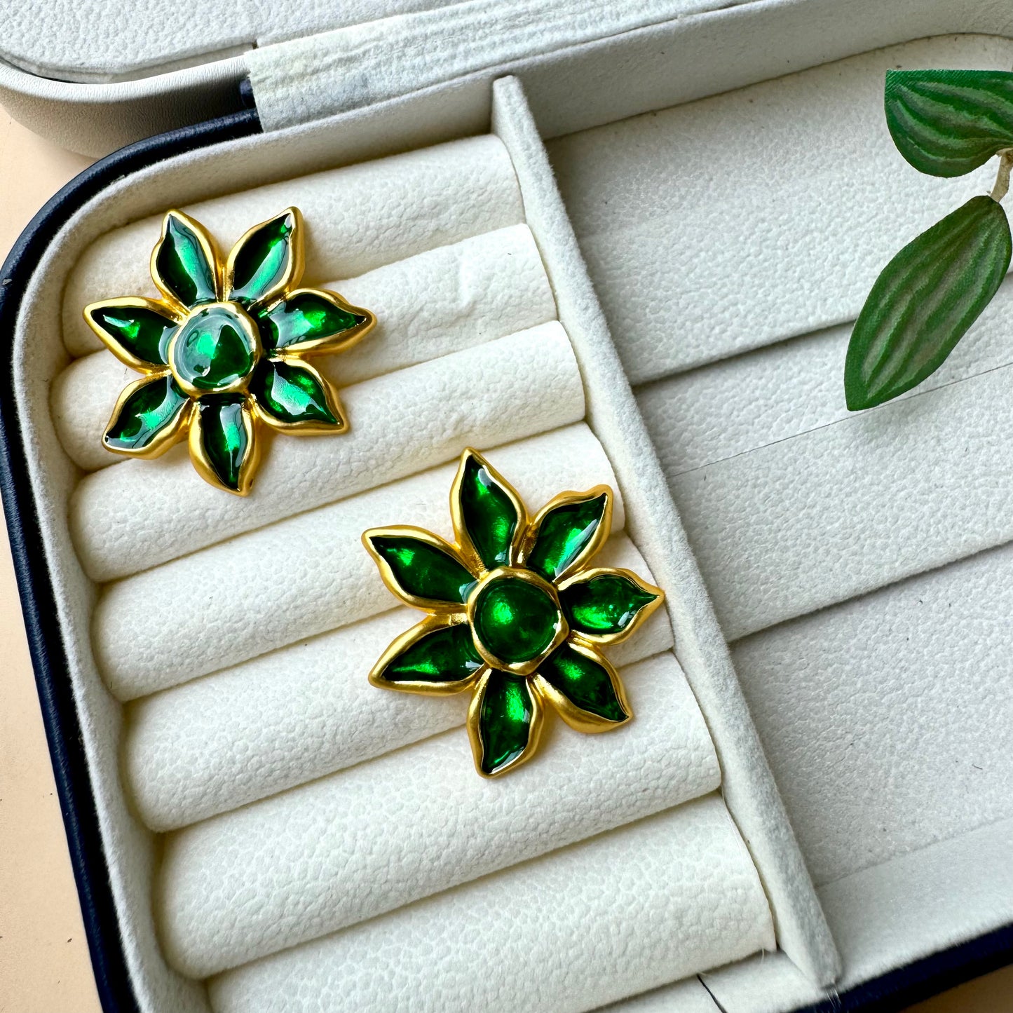 A pair of gold-plated floral earrings with vibrant green enamel petals, displayed on a white cushioned jewelry case. The earrings resemble blooming flowers with a glossy finish, adding an elegant and nature-inspired touch
