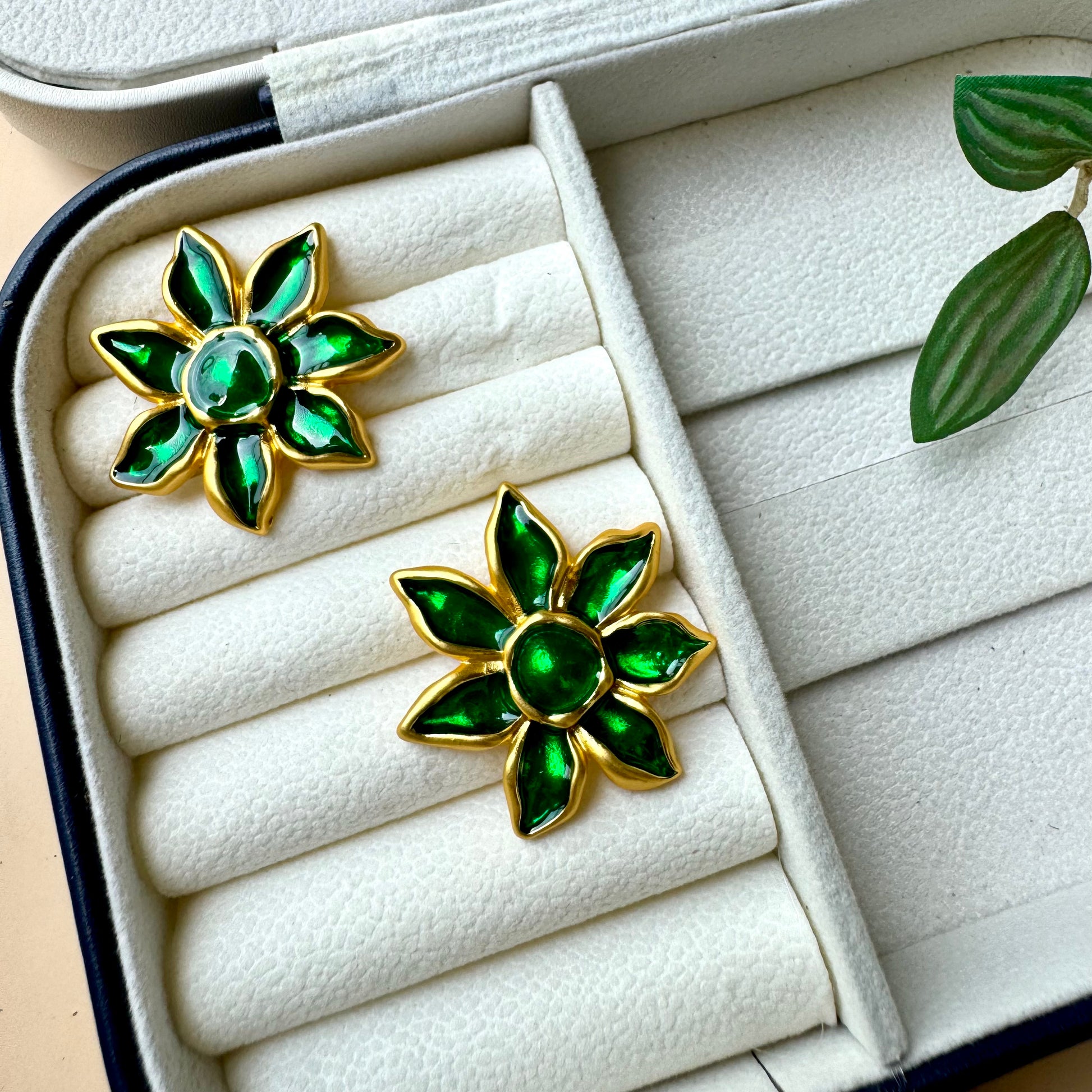 A pair of gold-plated floral earrings with vibrant green enamel petals, displayed on a white cushioned jewelry case. The earrings resemble blooming flowers with a glossy finish, adding an elegant and nature-inspired touch