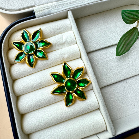 A pair of gold-plated floral earrings with vibrant green enamel petals, displayed on a white cushioned jewelry case. The earrings resemble blooming flowers with a glossy finish, adding an elegant and nature-inspired touch