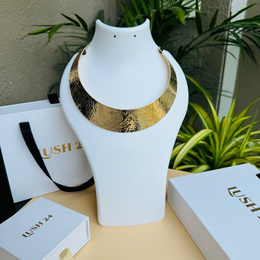 A dramatic gold metallic collar necklace with radiating spikes is displayed on a white mannequin bust. Lush 24 branded shopping bags and a jewelry box are visible in the background.