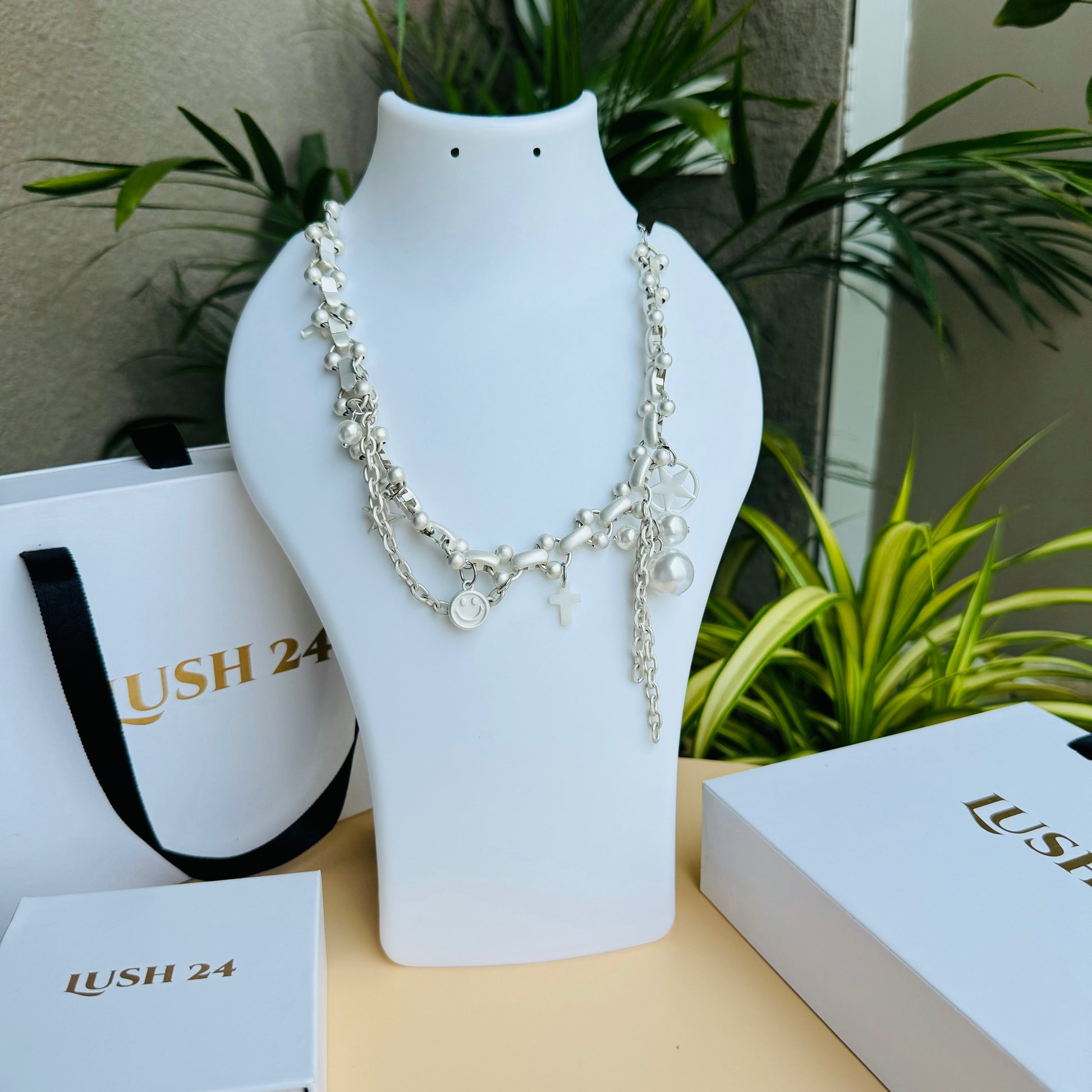 A silver charm necklace displayed on a white bust stand, featuring delicate chains adorned with small charms, including a smiley face, cross, pearl, and abstract shapes. The background includes Lush 24 branded packaging and green plants.
