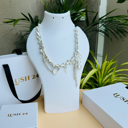 A silver charm necklace displayed on a white bust stand, featuring delicate chains adorned with small charms, including a smiley face, cross, pearl, and abstract shapes. The background includes Lush 24 branded packaging and green plants.