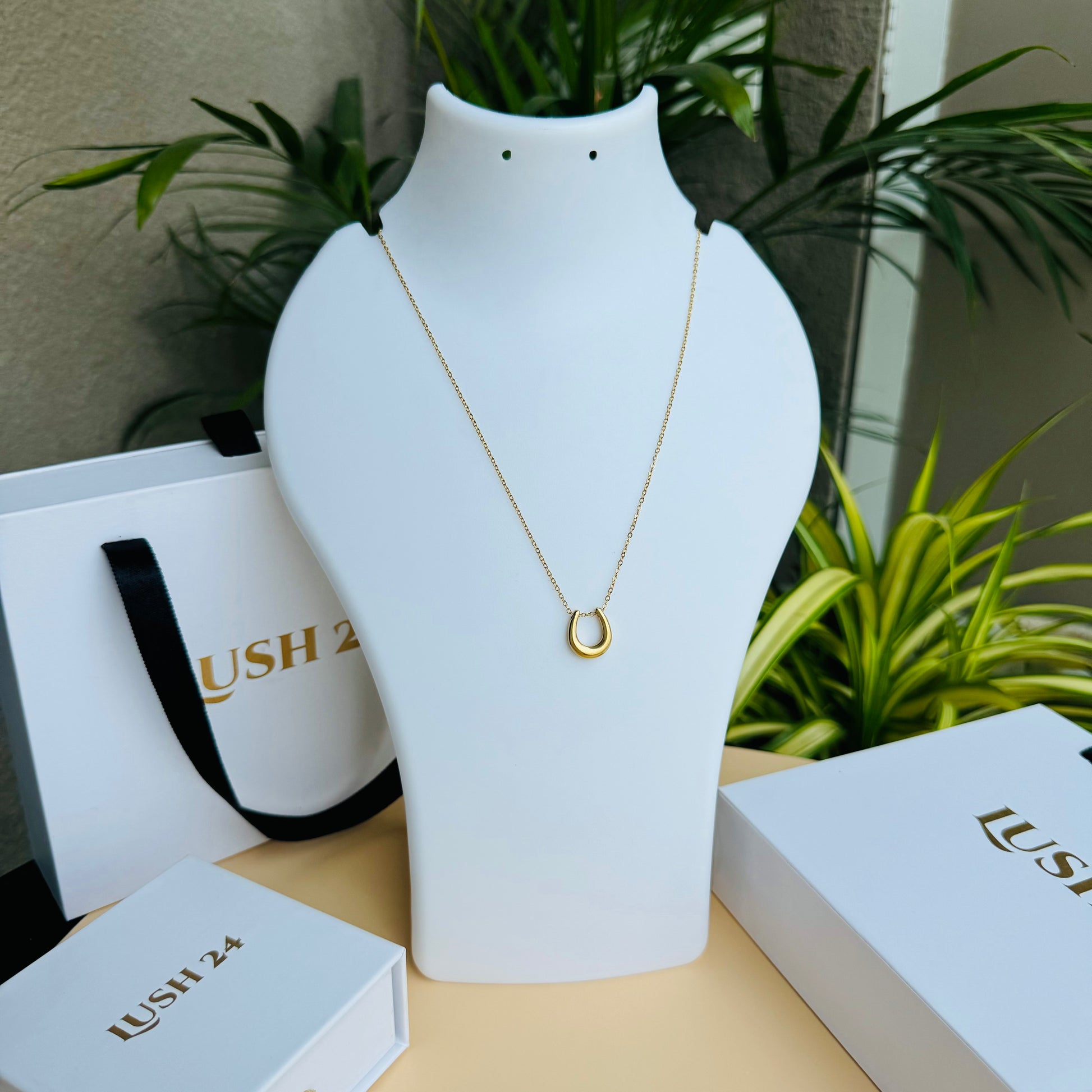 A delicate gold necklace featuring a minimalist horseshoe pendant, elegantly displayed on a white bust with LUSH 24 packaging in the background