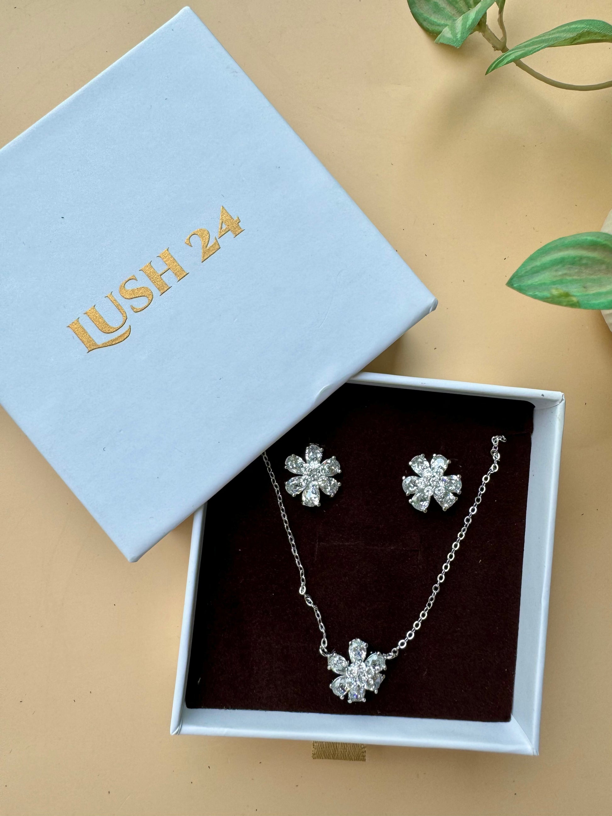 Elegant silver floral jewelry set featuring a delicate necklace with a matching pair of sparkling stud earrings, displayed on a textured beige tray with green leaf accents.