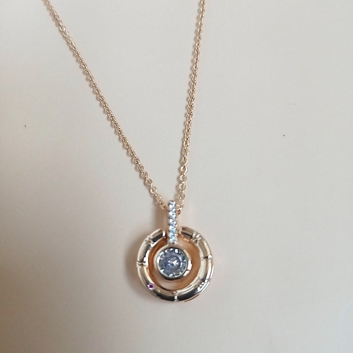Elegant rose gold necklace with a circular pendant featuring a central blue gemstone, accented with sparkling white stones on the bail, set against a neutral background