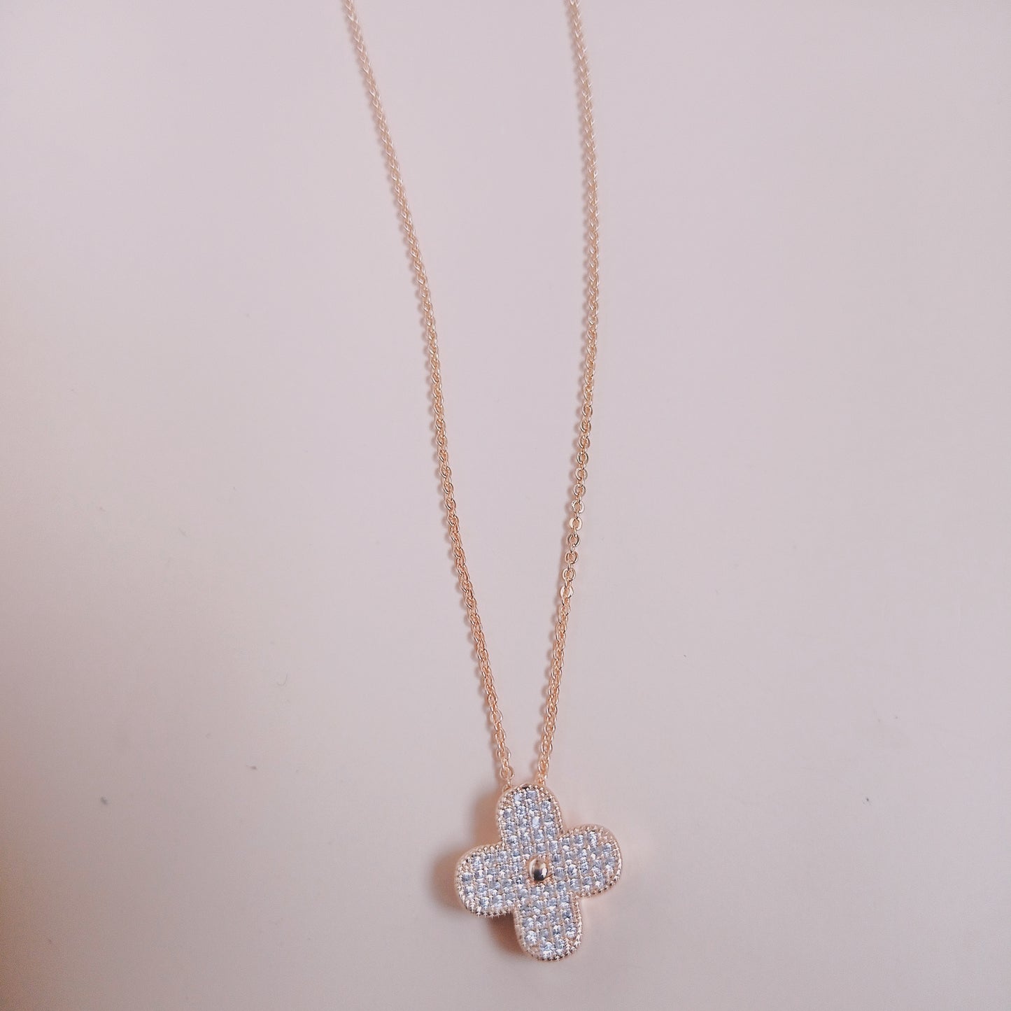 Elegant rose gold necklace with a four-leaf clover pendant encrusted with shimmering white stones, displayed against a soft pink background