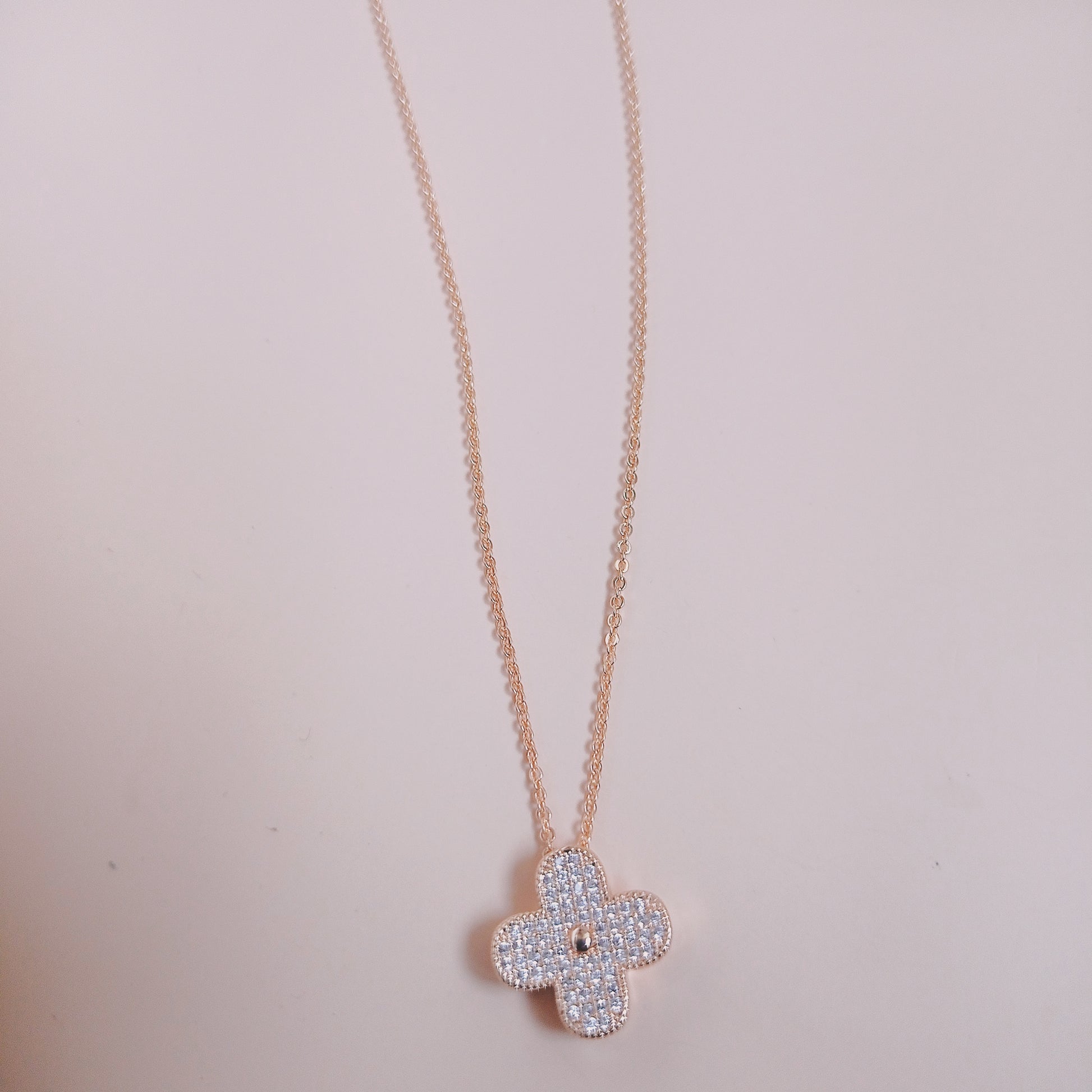 Elegant rose gold necklace with a four-leaf clover pendant encrusted with shimmering white stones, displayed against a soft pink background