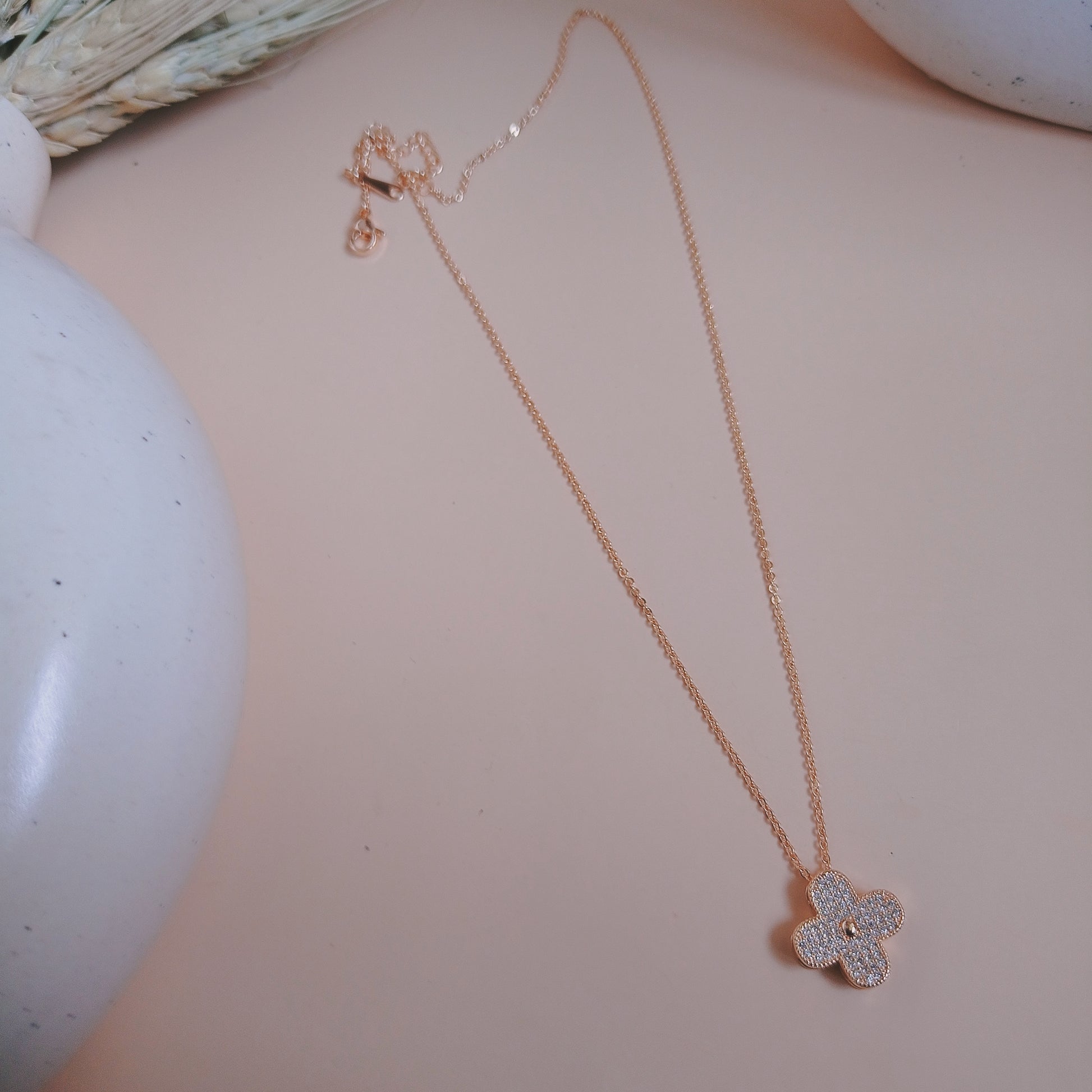 Elegant rose gold necklace with a four-leaf clover pendant encrusted with shimmering white stones, displayed against a soft pink background