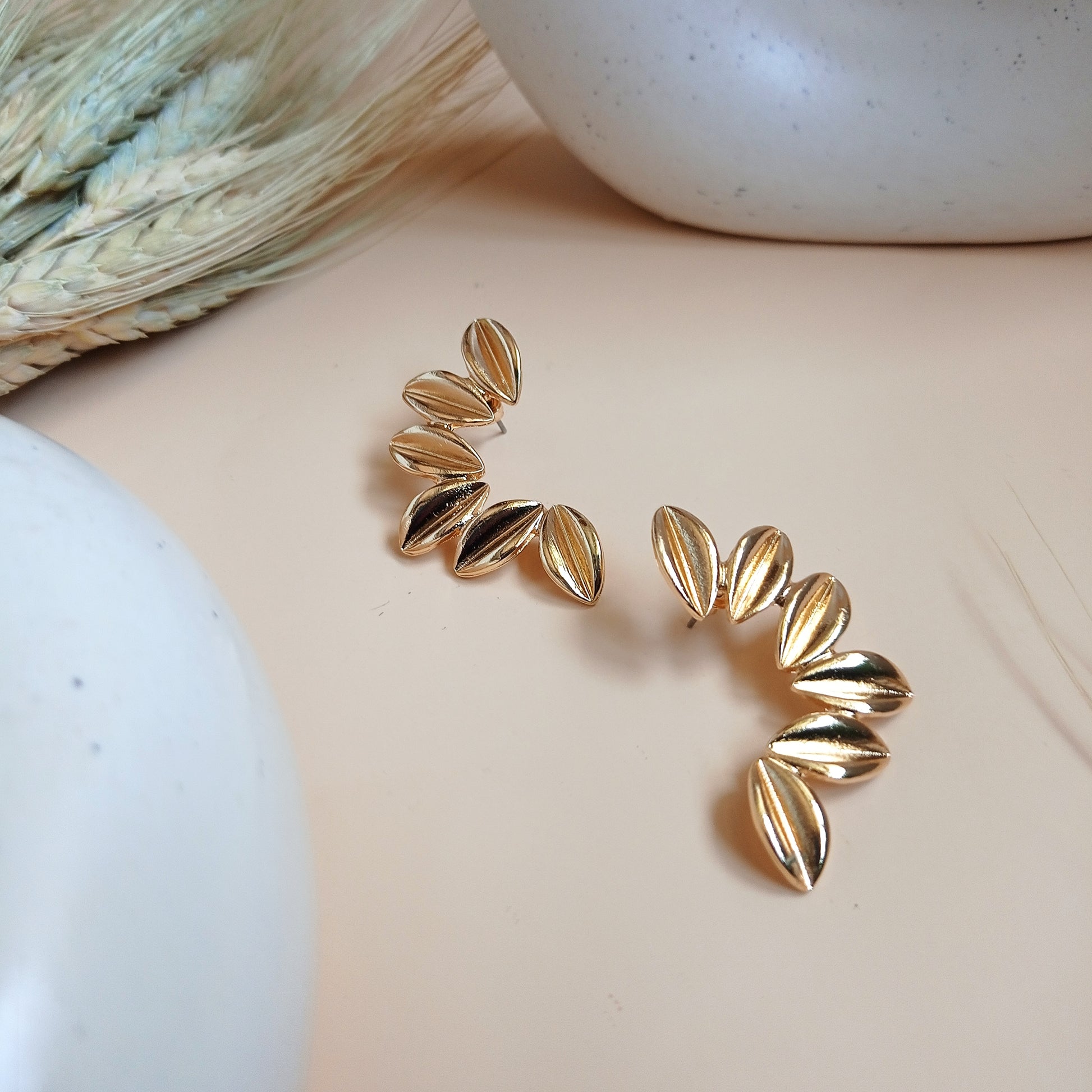 Unique gold statement earrings featuring a curved, leaf-inspired design with a brushed finish, elegantly displayed on a neutral beige background with ceramic and dried floral decor