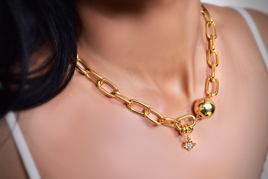 A close-up image of a woman's neck wearing a gold chain necklace with large, elongated links. The necklace features a polished gold sphere and a small dangling star-shaped charm adorned with a sparkling gemstone. The jewelry has a bold and elegant design, standing out against the woman's smooth skin