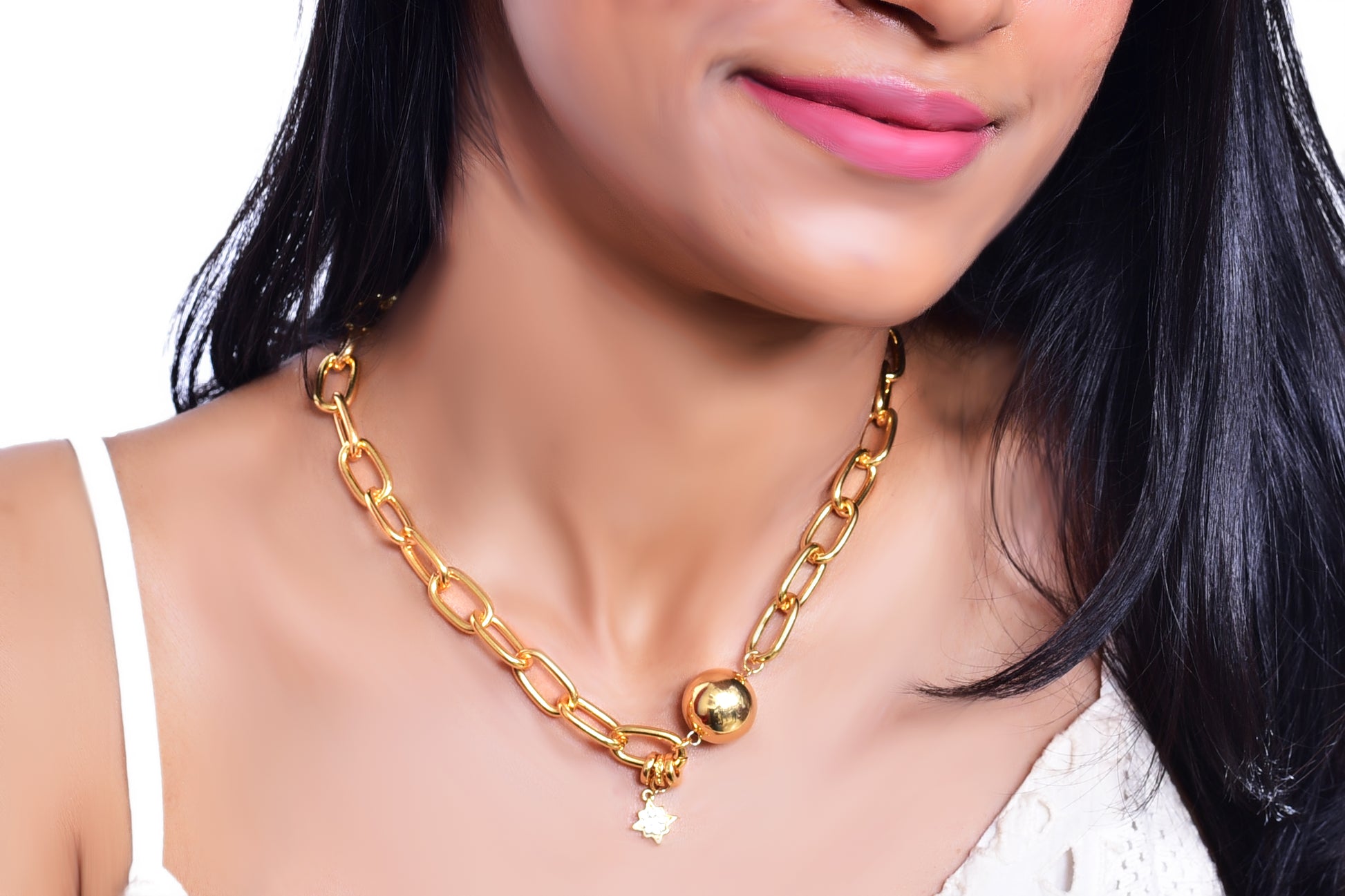 A close-up image of a woman's neck wearing a gold chain necklace with large, elongated links. The necklace features a polished gold sphere and a small dangling star-shaped charm adorned with a sparkling gemstone. The jewelry has a bold and elegant design, standing out against the woman's smooth skin