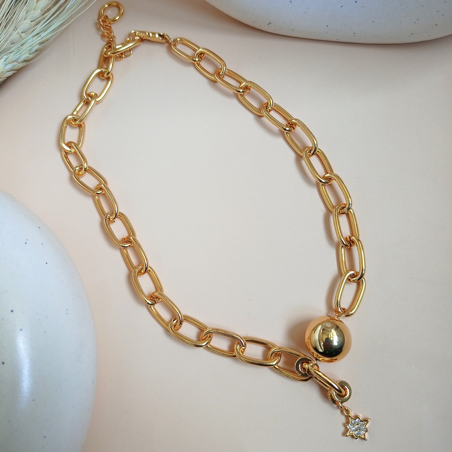 A close-up image of a woman's neck wearing a gold chain necklace with large, elongated links. The necklace features a polished gold sphere and a small dangling star-shaped charm adorned with a sparkling gemstone. The jewelry has a bold and elegant design, standing out against the woman's smooth skin