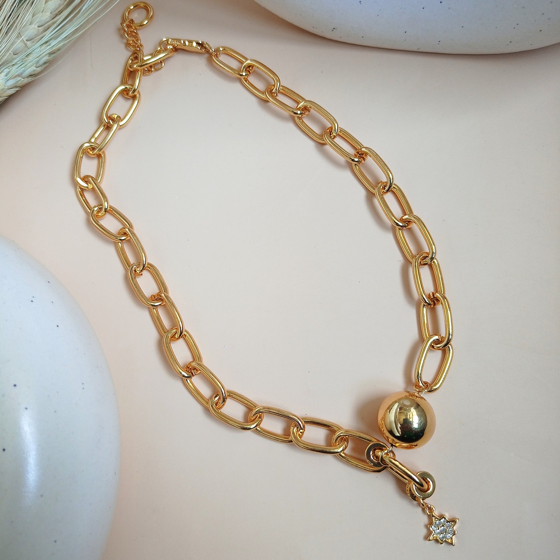 A close-up image of a woman's neck wearing a gold chain necklace with large, elongated links. The necklace features a polished gold sphere and a small dangling star-shaped charm adorned with a sparkling gemstone. The jewelry has a bold and elegant design, standing out against the woman's smooth skin