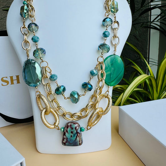 A statement multi-layered necklace featuring gold-tone chain links, faceted aqua beads, and large green gemstone accents. The design is completed with a unique marbled stone pendant, creating an elegant yet bold look. Displayed on a white jewelry bust with a tropical green backdrop.