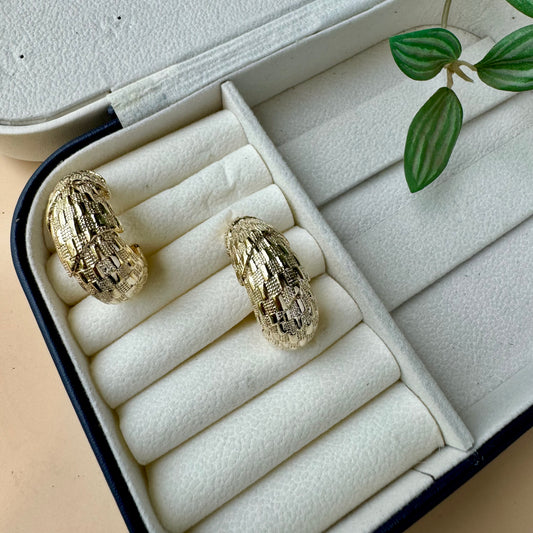 A pair of chunky, textured gold hoop earrings nestled in the soft, white velvet compartments of a dark blue and white jewelry case.