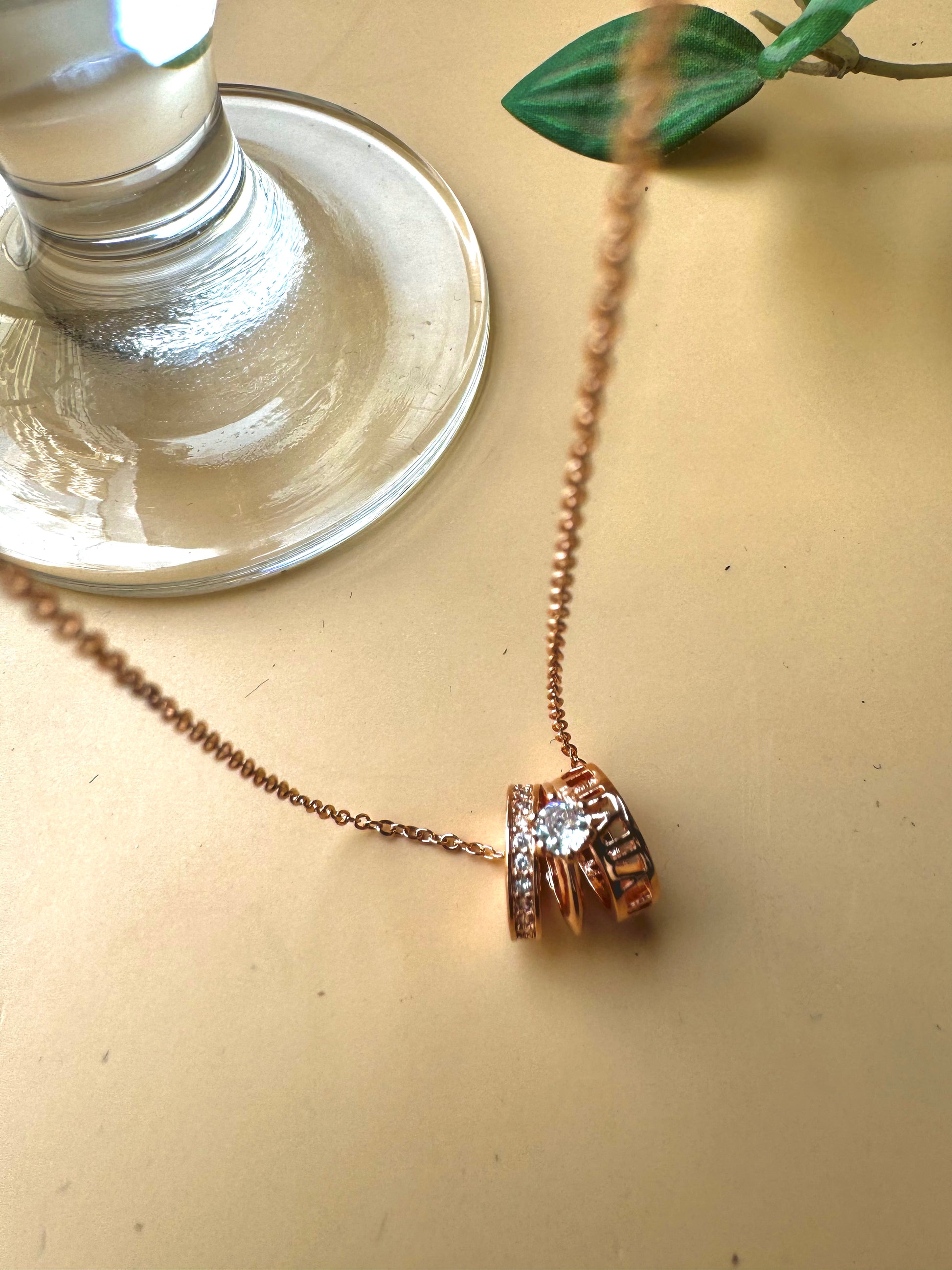 A delicate rose gold chain necklace featuring a pendant composed of stacked rings, one of which is set with clear gemstones resembling diamonds. The pendant has a contemporary, cylindrical design
