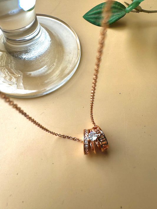 A delicate rose gold chain necklace featuring a pendant composed of stacked rings, one of which is set with clear gemstones resembling diamonds. The pendant has a contemporary, cylindrical design