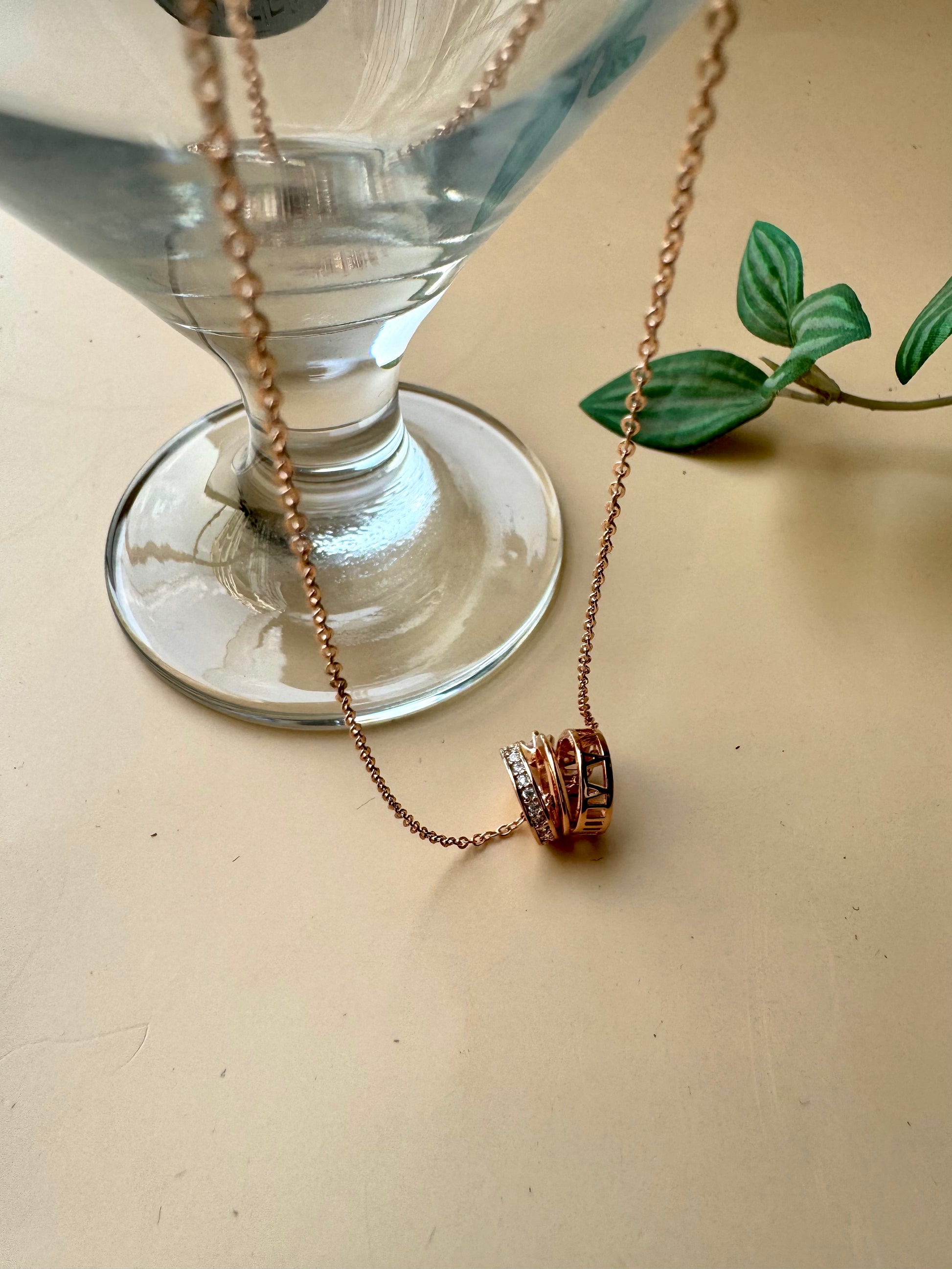 A delicate rose gold chain necklace featuring a pendant composed of stacked rings, one of which is set with clear gemstones resembling diamonds. The pendant has a contemporary, cylindrical design